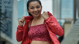 A photo of a smiling Filipino woman in a light red jacket and denim shorts, with buildings in the background